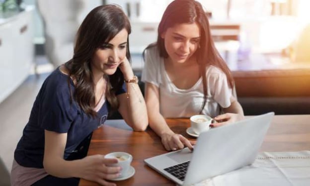 Two brunette women sitting at a table looking at a laptop screen together. They each have a cup of coffee. 
