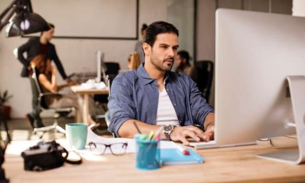 A man sitting at a desk looking at a monitor. On the desk is also a camera, a cup of pens, glasses, a mug, and papers. 