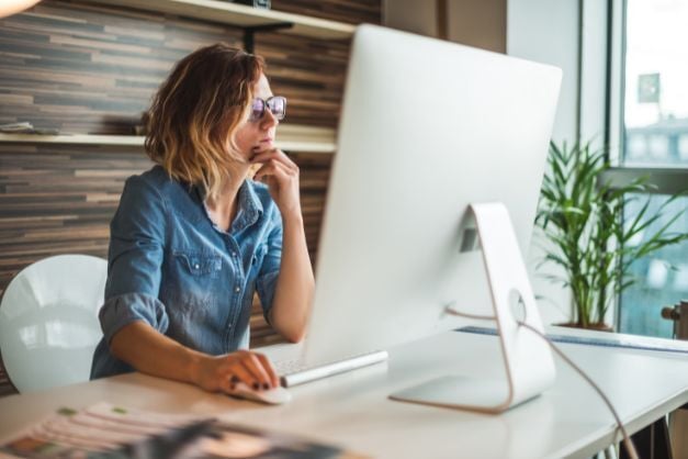 Woman seated at desk with large computer monitor in front of her, one hand on her chin, the other on her mouse, with a plant and window in background.