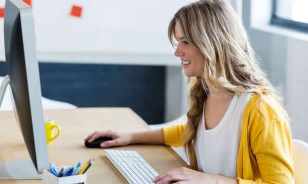 A woman sitting at a desk looking at a monitor screen smiling. She has one hand on the mouse and one on the keyboard. She is wearing a yellow sweater.