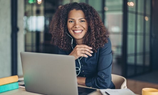An image of a woman with curly hair smiling at the camera. She has headphones in her ears and is seated in front of her laptop at a desk in an office with windows behind her.