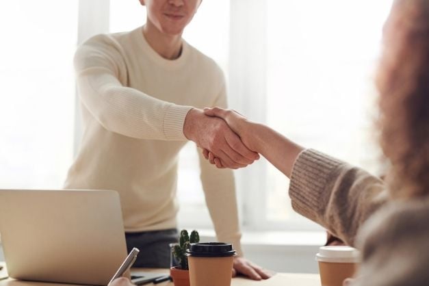 Two people, a man and a woman, shaking hands over a desk, one seated, one standing.