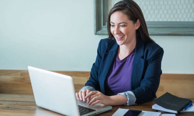 A woman wearing a blue blazer sitting at a table looking at a laptop screen. On the table are a cell phone and a notepad.