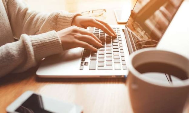 Hands resting on a laptop keyboard on a desk. Their sleeves are rolled up. There's a cell phone, glasses, and full mug of coffee on the desk. 