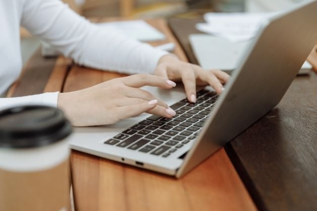 Woman's hands typing on laptop keyboard while seated at a desk; a coffee cup is visible in the foreground.