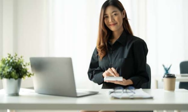 A woman is smiling while sitting at a desk with a laptop, a notebook, and glasses on the desk with a coffee cup beside them.