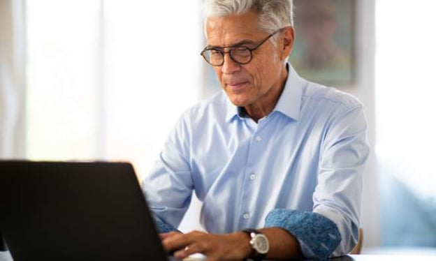 An older man with glasses sits at a table, looking at a laptop screen. He is wearing a blue dress shirt and a watch. A window is in the background.