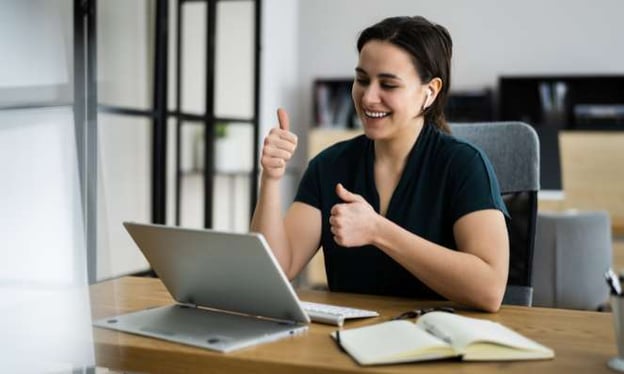 A woman sitting at a desk in a black shirt smiles at the laptop screen, giving both thumbs up to the screen. Beside the laptop is a notebook.