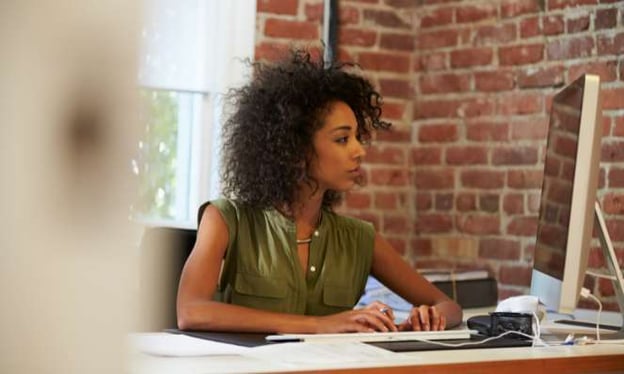 Woman sitting at a desk looking at a computer monitor. The desk has papers all over it. The woman has her hands rested on the keyboard.