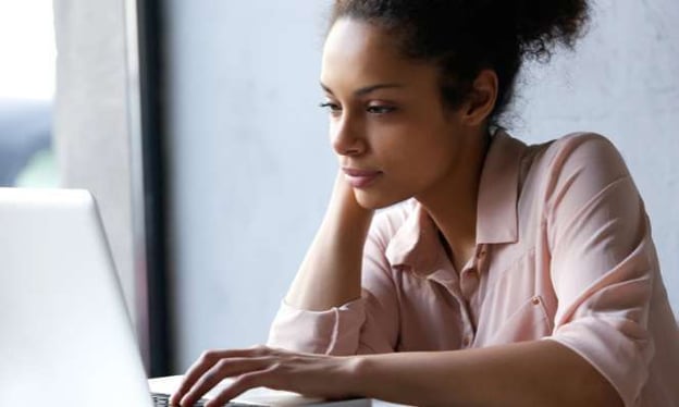 A woman is looking at a laptop screen, wearing a pink dress shirt and her hair in a bun.