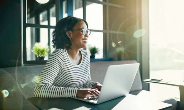 A woman is smiling, sitting at a desk, and wearing glasses. On the desk is an open laptop. To her left is the sun shining through the window.