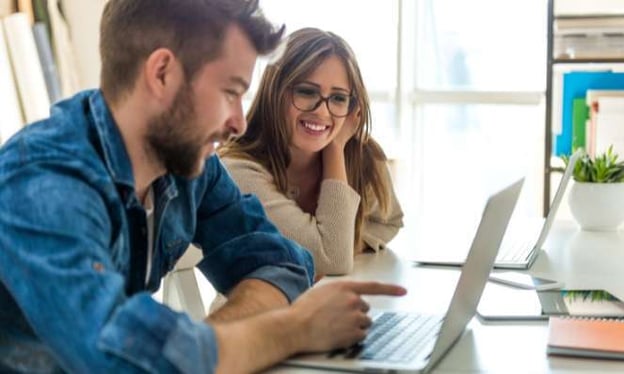 A man and a woman sit at a desk, smiling and looking at a laptop screen. On the desk is another laptop in front of the woman, as well as a notebook.