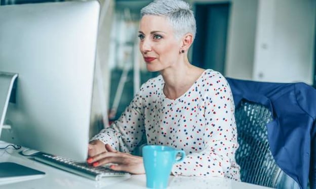 A woman sits at a desk working on a desktop, looking at a monitor screen, with her hands resting on a keyboard. She has a coffee cup next to her.