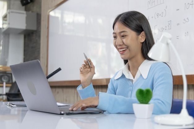Woman seated at desk, one hand on her laptop keyboard, one hand in the air, holding a pen, with a lamp in the foreground and whiteboard in the back.