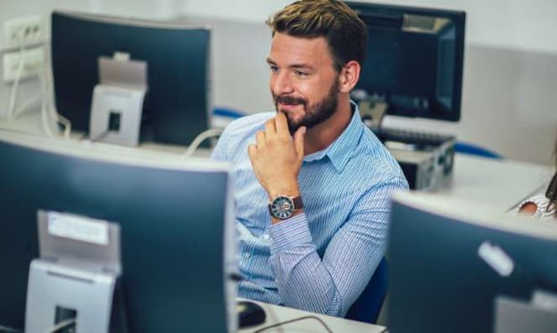 A man sitting at a desk looking at a monitor screen. He is wearing a blue dress shirt and a watch with his chin resting on his hand. 