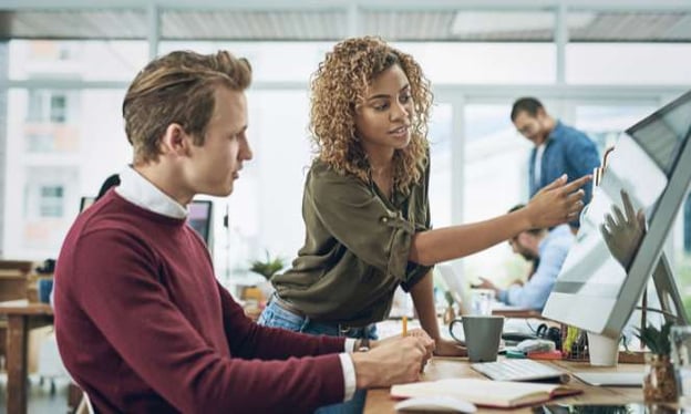 A man sits at a desk working on a monitor. There is a woman standing next to him, pointing at the monitor screen. 