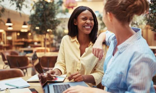 Two women at a restaurant are sitting at a table, smiling at one another. On the desk in front of them is a laptop and notebook.