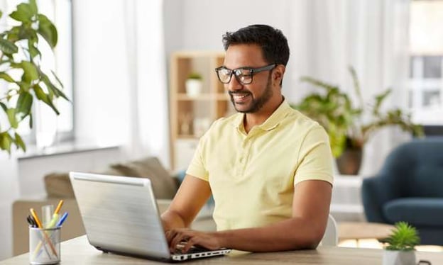 A man wearing a yellow t-shirt and black glasses is sitting at a desk and working on a laptop while smiling. On the desk is a small plant and pens. 
