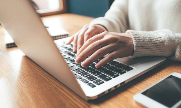 Hands resting on a laptop keyboard while seated at a desk. The laptop is on the desk next to a cell phone and notebook. Window in background.