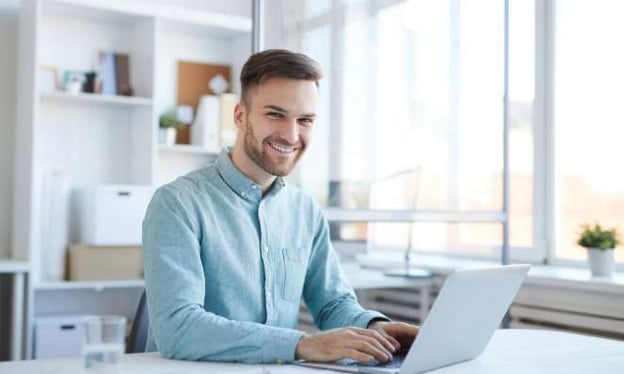 A man is sitting at a desk working on a laptop while smiling. He is wearing a blue dress shirt. In the background are shelves with books and baskets. 