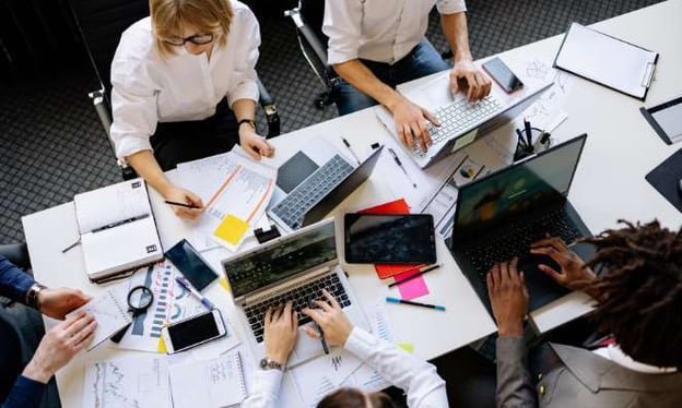 A group of people are working on their laptops while sitting around a shared table. There is one woman and four men.