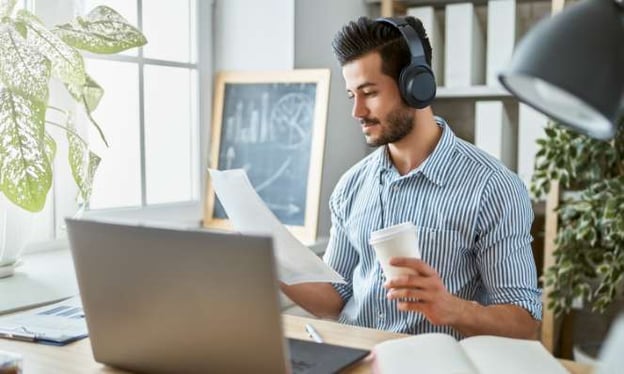 A man sits at a desk with headphones on with a coffee cup in one hand, and papers in the other hand. In front of him is an open laptop. 