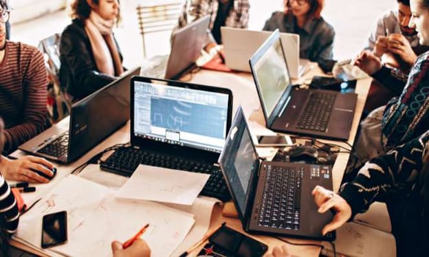 A group of eight people are sitting around a table while working on their laptops.
