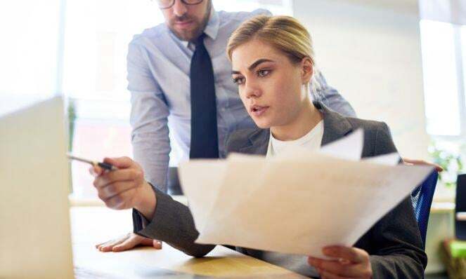 Woman working on her computer while holding a bunch of papers. A man stands behind her looking at the monitor as she points to it holding a pen.