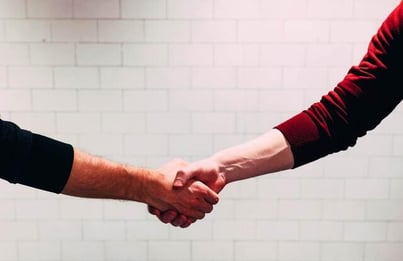 Two men's arms shaking hands in front of a white brick wall. The arm on the right is wearing a red shirt; the arm on the left is wearing black.