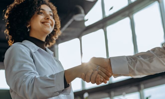 A smiling woman shaking hands with another person whose hand and arm are only visible in the photo. There is a wall of windows behind them.